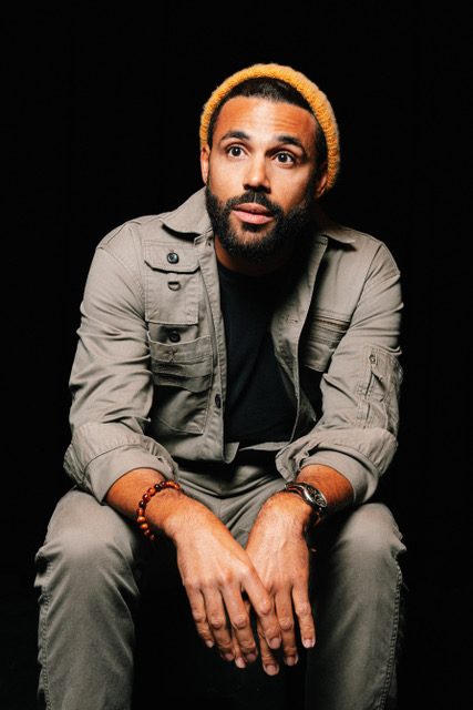 Seated portrait in a mustard beanie and olive jacket against black