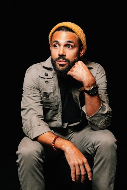 Seated, chin on hand, in a mustard beanie against black