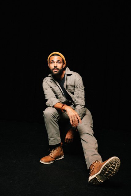 Full-length seated portrait, one boot forward, against black